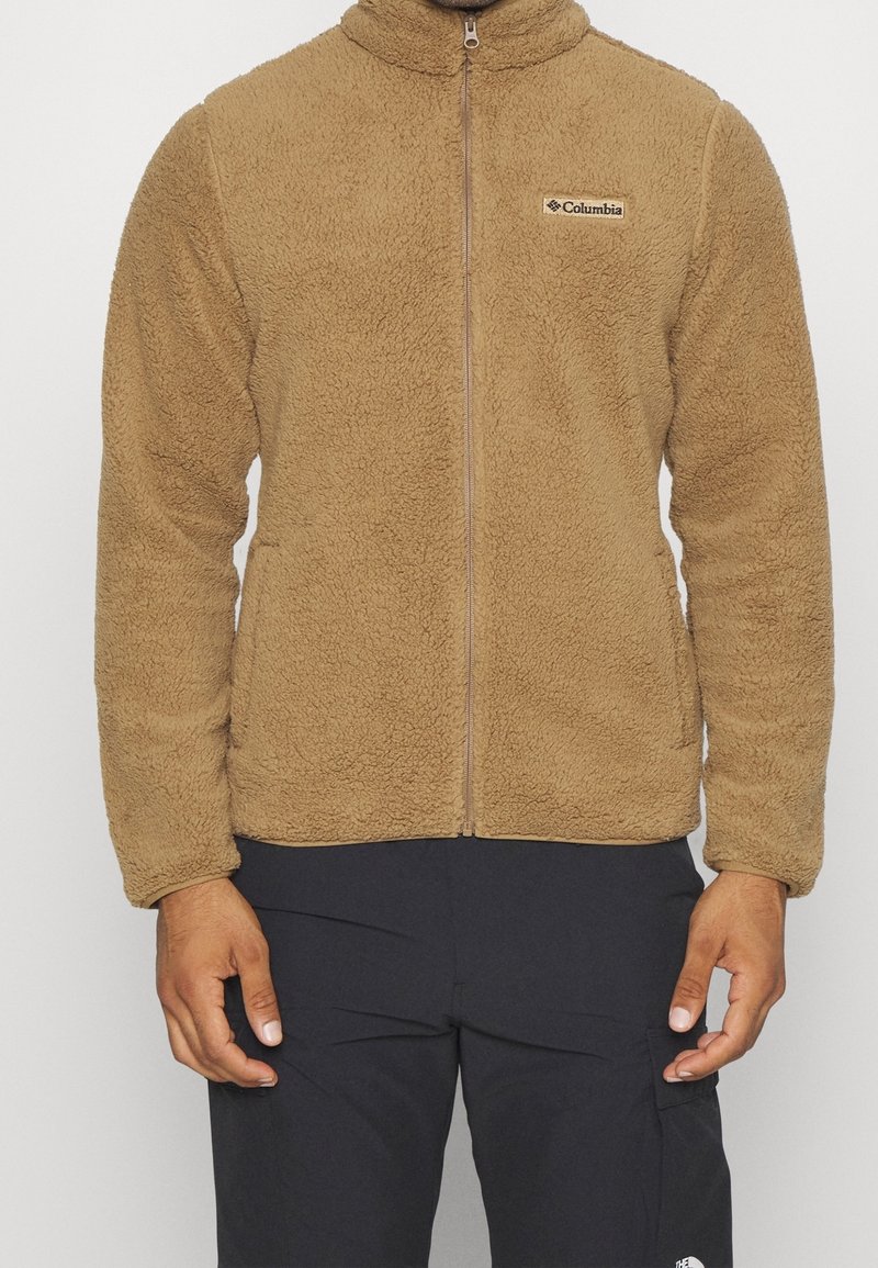 Brun fleecjacka med hög krage, framzipper och logopatch på vänster bröst. Tyget har en strukturerad, mjuk yta.