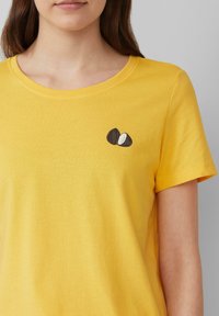 T-shirt en coton jaune avec un col rond, ornée d'un petit design brodé de graines brunes et blanches sur le côté gauche.