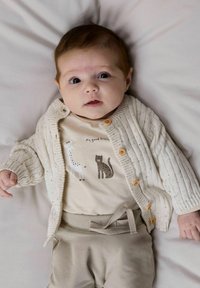 Cardigan crème clair à texture côtelée, fermeture par boutons. Body beige avec des motifs d'animaux. Pantalon beige doux avec taille nouée.