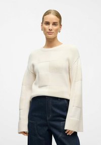 Jersey color crema con un patrón texturizado de grandes cuadros, mangas anchas y un cuello redondo, combinado con jeans oscuros.