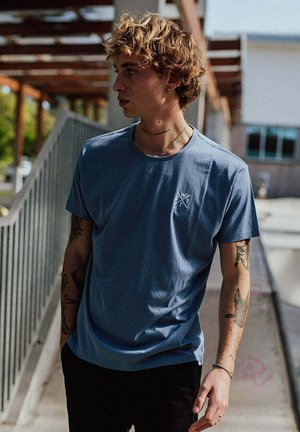 Jong persoon met krullend haar, gekleed in een blauw T-shirt en zwarte broek, staand buiten bij een reling met zichtbare tatoeages op de armen.