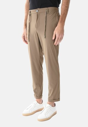 Uomo che indossa pantaloni beige slim fit con polsini alla caviglia e sneakers bianche, in piedi davanti a uno sfondo semplice.
