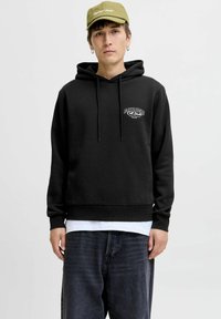 Sudadera negra con cordones y detalle del logo en el frente, llevada sobre una camisa blanca. Combinada con pantalones vaqueros oscuros de corte holgado y una gorra verde claro.