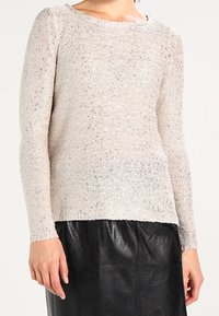 Suéter de punto de color beige claro con mangas largas, material texturizado y sutiles toques de brillo, combinado con una falda de cuero negra.