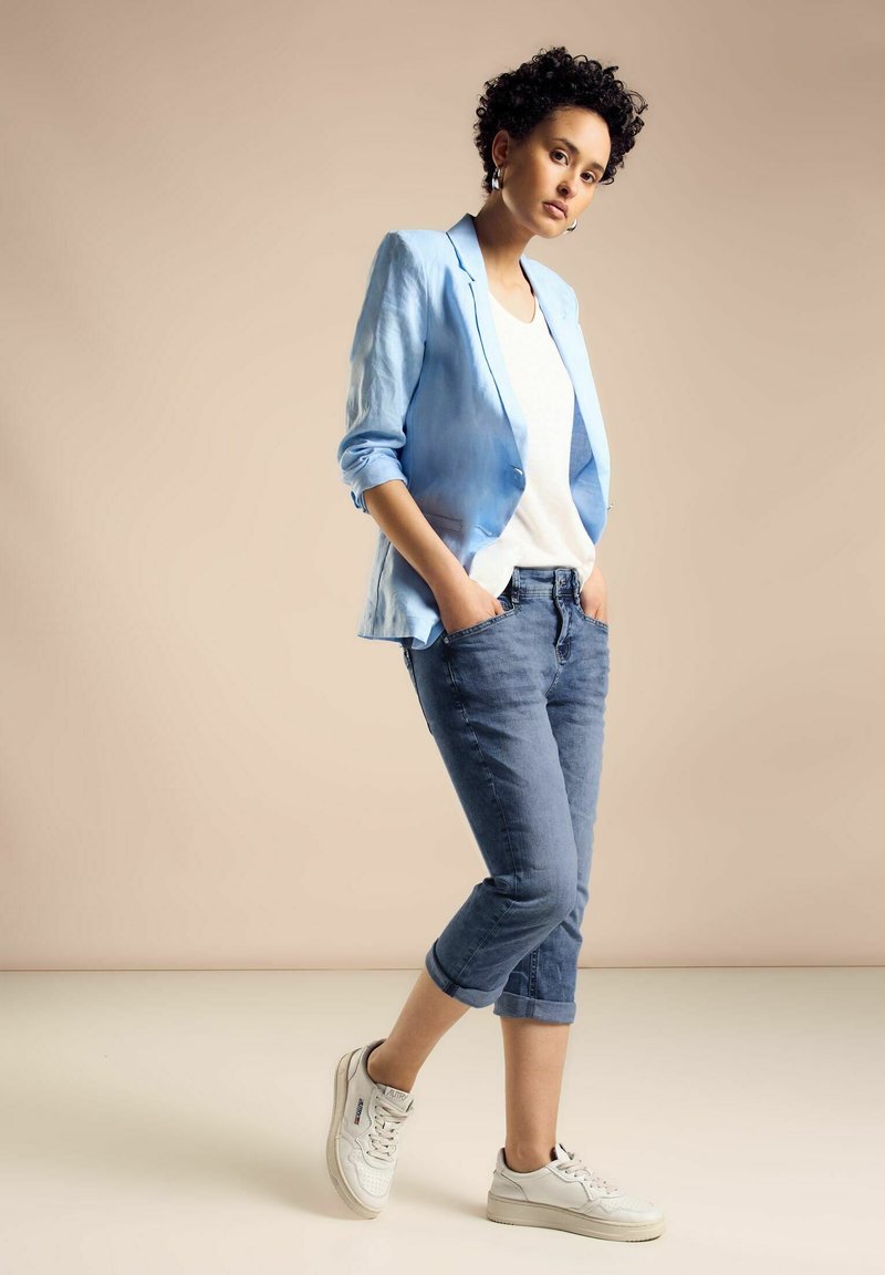 Hellblaues Leinenblazer über einem weißen T-Shirt, kombiniert mit hochgekrempelten blauen Jeans und weißen Sneakers. Strukturierter Stoff und lässiger Schnitt.