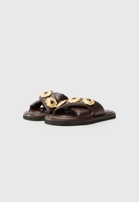 Brown leather sandals with cross straps, featuring gold floral accents and dark round beads. Flat sole with minimal tread design.