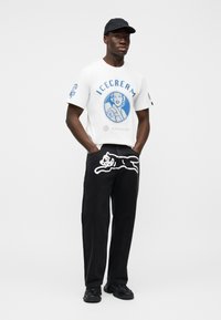 Λευκό γραφικό T-shirt με μπλε κείμενο "ICECREAM" και εικόνα. Μαύρο παντελόνι με λευκό σχέδιο γάτας. Μαύρο καπέλο και χοντρά μαύρα παπούτσια.