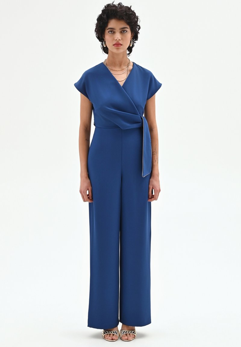 Blauwe jumpsuit met een overslagontwerp, korte mouwen en wijde pijpen. Gemaakt van gladde stof met een strikdetail in de taille, gecombineerd met sandalen met bandjes.