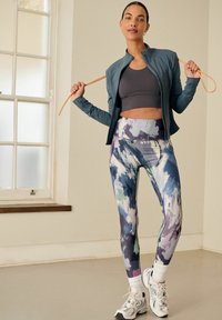 Letvægts, lynlåset teal jakke over en grå sports-bh, parret med farverige, abstrakte mønstrede leggings i blå, hvid og grøn. Hvide sneakers.