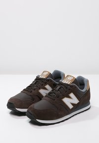 Baskets en daim marron avec des accents gris, un logo blanc et une languette rembourrée. Elles présentent une semelle en caoutchouc noire et un design à lacets.