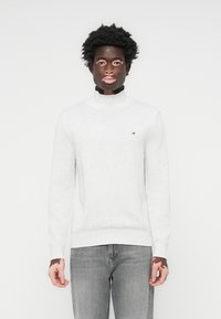 Lysegrå turtleneck genser laget av myk strikket stoff. Har ribbestrikkede mansjetter og nederkant. Lite logo på venstre bryst. Sammen med grå jeans.