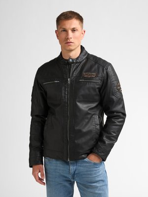 Mann trägt eine schwarze Lederjacke mit Reißverschlusstaschen und Aufnähern, steht mit einer Hand in der Tasche der blauen Jeans vor einfachem Hintergrund.