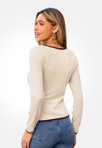 Maglia beige a maniche lunghe con costine, con un collo e orlo a contrasto marrone. Abbinata a jeans blu a vita alta, con un design aderente.