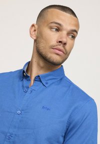 Chemise bleu à boutons en tissu léger, dotée d'un col pointu, d'un logo brodé sur la poitrine et de boutons assortis.