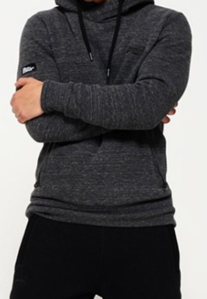 Persoon die een donkergrijze hoodie draagt met voorzakken en trekkoorden, armen over elkaar op de borst, zittend een zwarte broek te dragen.