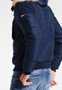 Man i marinblå jacka med pälsmössa och ljusblå jeans, med händerna vilande nära bakfickorna, vänd något bort.