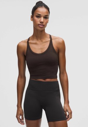 Mujer con cabello oscuro recogido en un moño alto, vistiendo un top corto marrón oscuro y pantalones cortos deportivos negros de talle alto contra un fondo liso.