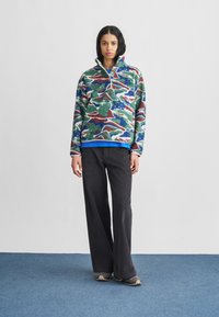 Πουλόβερ φλις με πολύχρωμο σχέδιο που περιλαμβάνει σπείρες σε πράσινο, μπλε και μαρόν χρώμα· λαιμόκοψη με φερμουάρ· συνδυασμένο με μαύρο παντελόνι φαρδιάς γραμμής και γκρι παπούτσια.