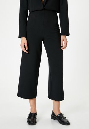 Svarta vida byxor i ett mjukt tyg, med hög midja och veckdetaljer, kombinerat med svarta loafers för en enhetlig look.