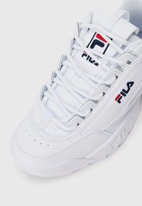 Sneaker FILA blanche avec des lacets épais, une pointe perforée et un logo brodé sur le côté et la languette, inclinée contre un fond blanc.