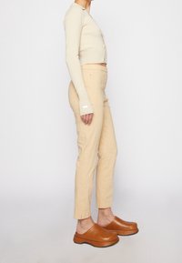 Femme portant un cardigan court crème, un pantalon beige taille haute et des chaussures compensées en cuir brun, se tenant de profil sur fond blanc.