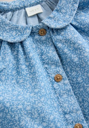 Sininen kukkakuviollinen paita, jossa pyöreä kaulus, nappikiinnitys ja teksturoitu kangas. Napit ovat vaalean puun värisiä ja niissä on ympyrämäinen kuvio.