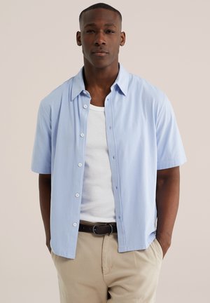 Junger Mann trägt ein hellblaues, kurzärmliges Hemd über einem weißen Tank-Top und beige Hosen, steht mit den Händen in den Taschen vor einem einfarbigen Hintergrund.