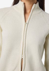Pull en tricot côtelé crème avec zip et col montant, mettant en valeur un tissu texturé et une coupe ajustée.