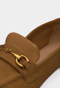 Mocassin en daim marron avec un détail en métal doré en forme de mors de cheval sur le dessus et un design à bout rond.