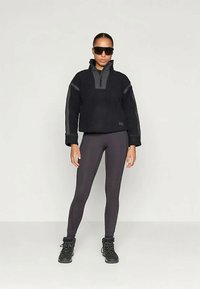 Donna che indossa un pullover nero in pile, leggings grigio scuro, scarponi da trekking neri e occhiali da sole neri di grandi dimensioni, in piedi contro uno sfondo neutro.