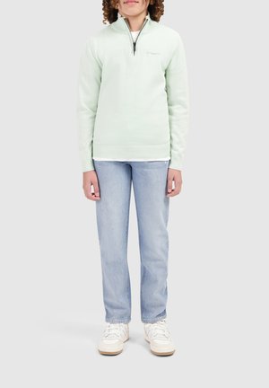 Hellgrüner, gestrickter Pullover mit einem Reißverschlusskragen, kombiniert mit hellblauen, geraden Jeans und weißen Sneakern.