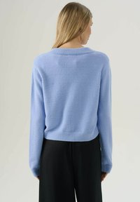 Una donna con lunghi capelli biondi indossa un maglione morbido blu chiaro e pantaloni neri, rivolta verso l'esterno su uno sfondo chiaro e semplice.