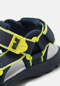 Marinblå sandal med limegröna detaljer, med justerbara remmar, en texturerad svart sula och ett tassavtryck som logotyp på spännet.