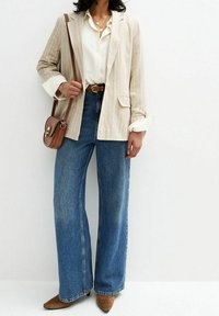 Nainen, jolla on päällä kerma valkoinen bleiseri, valkoinen paita, leveälahkeiset siniset farkut, ruskea vyö, ruskeat teräväkärkiset kengät ja ruskea olkalaukku, seisoo valkoista seinää vasten.