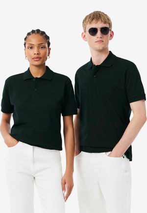 Deux personnes portant des polos noirs et des pantalons blancs, l'une avec des cheveux tressés et l'autre portant des lunettes de soleil, toutes deux les mains dans les poches.
