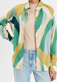 Personne portant une chemise à boutons de motif abstrait multicolore sur un top côtelé beige et un jean bleu clair, la main reposant sur la hanche.