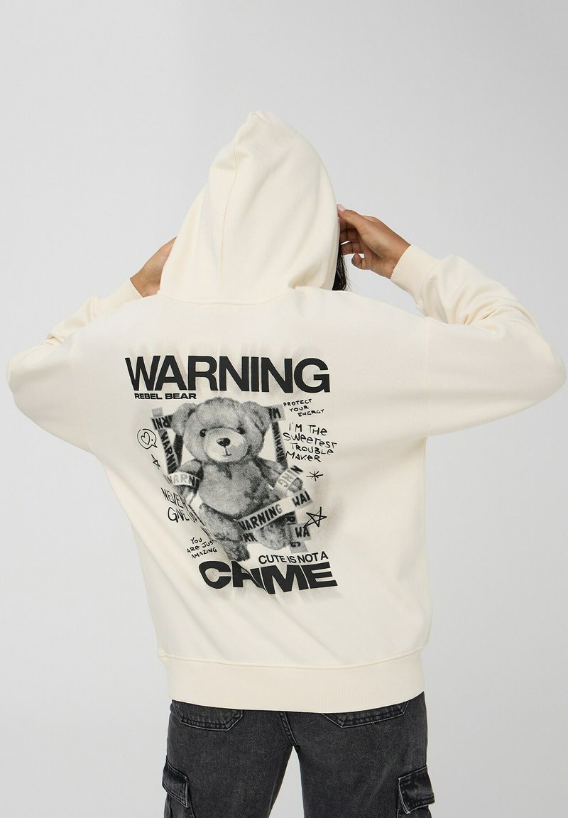 Kremowa bluza z kapturem z dużą czarną grafiką przedstawiającą niedźwiedzia i dekoracyjnym napisem, podkreślona sznurkiem w kapturze i ściągaczami przy mankietach.