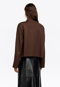 Brązowa krótka bluza z wysokim kołnierzem, luźnym krojem i opuszczonymi ramionami, zestawiona z błyszczącą czarną skórzaną spódnicą.