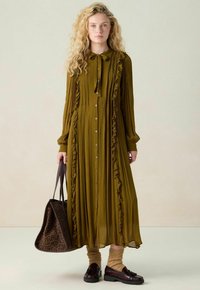 Robe longue olive avec un devant à boutons, des détails en volants et un nœud à col. Assortie avec un sac fourre-tout à motifs et des chaussettes marron.