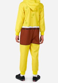 Mono amarillo con capucha, con una textura similar a la esponja y acentos circulares. Sección marrón en la cintura con líneas en negro y blanco.