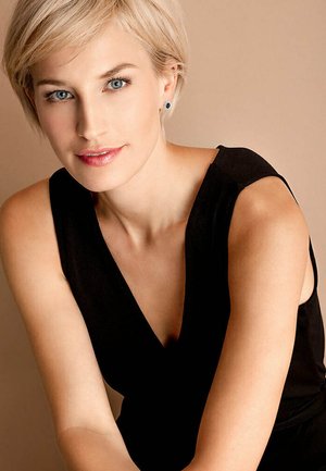 Model trägt ein ärmelloses schwarzes Kleid mit tiefem V-Ausschnitt, kurze blonde Haare, blaue Augen und Steckerohrringe vor einem neutralen Hintergrund.