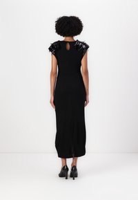 Robe noire sans manches avec des accents de franges sur les épaules, longueur genoux et une ouverture en trou de serrure au dos, portée avec des chaussures à talons noires.
