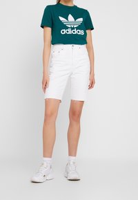 Turkusowa bawełniana koszulka z białym logo Adidas, zestawiona z białymi szortami z wysokim stanem o surowym wykończeniu. Uzupełniona białymi sneakersami.