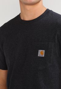 T-shirt in cotone grigio scuro con scollo rotondo e tasca sul petto a sinistra, caratterizzata da un'etichetta quadrata del logo Carhartt arancione e bianco.