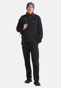 Man in een blauwe beanie, zwarte fleece met kwartrits, zwarte broek en wandelschoenen, die een zwarte rugzak op één schouder draagt.