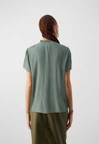 Kurzärmlige, olivgrüne Bluse mit strukturiertem Muster und Rüschenausschnitt, kombiniert mit einem langen, passenden Rock.