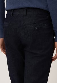 Pantalones de mezclilla oscura con una textura suave, que cuentan con bolsillos traseros con cierres de botón y un corte a medida. Llevados con un suéter azul.