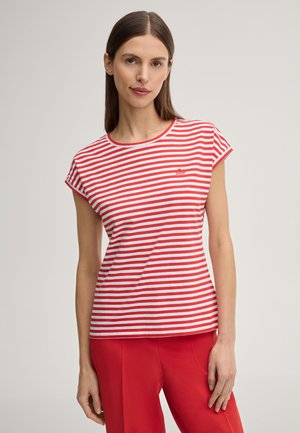 Vrouw met steil bruin haar draagt een rood-wit gestreept shirt met korte mouwen en een rode broek, staand tegen een effen lichte achtergrond.