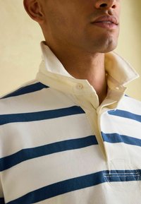 Polo w paski w kolorze granatowym i białym, z zapięciem na guziki przy kołnierzyku oraz teksturowanym materiałem z przeszytymi akcentami.