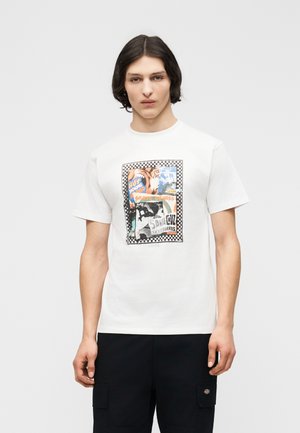 Młody mężczyzna z ciemnymi włosami do ramion, ubrany w białą koszulkę z nadrukiem deskorolki i czarne spodnie, na jednolitym tle.